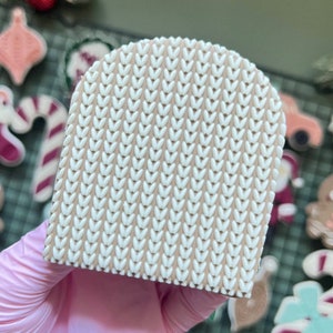 Op de afbeelding: Een beige rechthoekig object met een wit breipatroon. Het object wordt vastgehouden in een roze gehandschoende hand. Kerstkoekjes staan op de achtergrond, waaronder een peperkoekmannetje en zuurstokken.