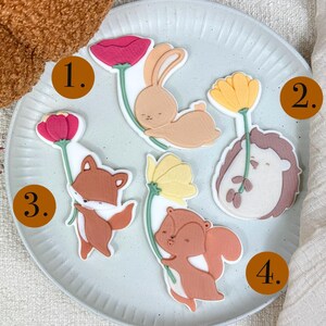 Puede incluir: Una colección de decoraciones caprichosas con temas de animales, que incluyen un conejo, un erizo, un zorro y una ardilla, cada uno sosteniendo una flor. Las decoraciones son de colores pastel suaves, dispuestas sobre un plato azul claro. El tema general es la naturaleza y la primavera.
