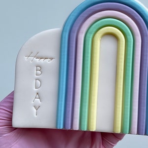 Könnte beinhalten: Ein weißer Tortendekor mit einem Regenbogenbogen und den Worten "Happy BDAY" auf der Seite.
