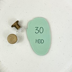 Könnte beinhalten: Ein weißer Keramikteller mit einer grünen, unregelmäßig geformten Karte, auf der "30 HBD" steht. Auf dem Teller befindet sich eine kleine goldene Stecknadel.