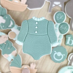May include: A collection of baby shower cookies decorated with green and brown icing. The cookies feature various baby-themed designs, including a baby onesie, a baby bottle, a baby carriage, and a baby rattle. The cookies are arranged on a wooden platter.