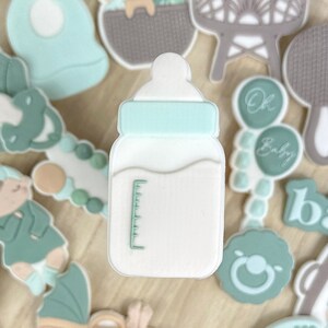 Peut inclure: Une collection de décorations sur le thème de bébé dans des tons de vert, blanc et beige, disposées sur un plateau en bois. Les décorations comprennent des biberons, des vêtements de bébé, des hochets et le mot "baby".