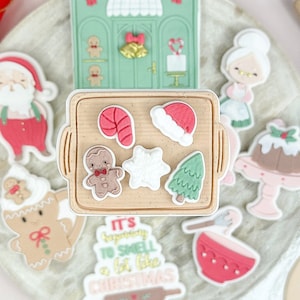 Puede incluir: Un juego de cortadores de galletas de silicona con temática navideña, que incluye una casa de jengibre, un hombre de jengibre, un bastón de caramelo, un árbol de Navidad, un copo de nieve, un gorro de Papá Noel y un tazón de pudín. Los cortadores están sobre una superficie de mármol con un rodillo y una bolsa de harina.