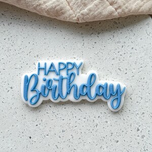 Puede incluir: Un adorno para pastel blanco con las palabras "Happy Birthday" en letras azules.