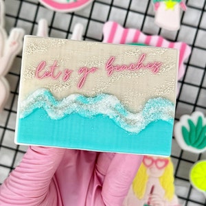 Könnte beinhalten: Ein rechteckiger Keks mit einer Strandszene. Er hat eine hellbraune Oberseite mit rosa Text "let's go beaching". Darunter befindet sich ein blauer Ozean mit weißen Wellen. Weitere Kekse zum Thema Strand sind im Hintergrund.
