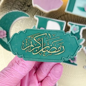 Könnte beinhalten: Ein grün und gold dekorierter Keks mit arabischer Kalligraphie, die "Ramadan Kareem" lautet. Der Keks wird von einer behandschuhten Hand gehalten.