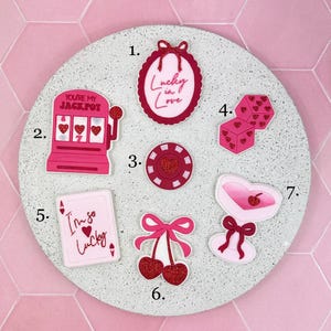 Puede incluir: Una colección de amuletos rosas y rojos con temática de casino, que incluyen dados, una carta de juego y una máquina tragamonedas. Los amuletos están dispuestos sobre una superficie redonda de color gris claro. Algunos amuletos tienen el texto "Lucky in Love" y "I'm so Lucky".