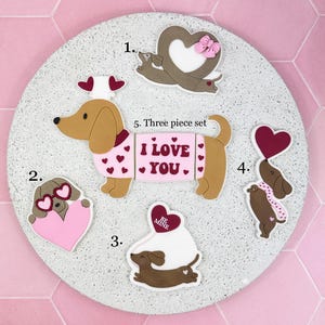 Puede incluir: Un juego de cinco piezas de decoraciones de perros salchicha con temática de San Valentín. El juego incluye varios diseños de perros con corazones y la frase "I Love You". Las decoraciones son en tonos marrón, rosa y blanco, con un fondo rosa claro.