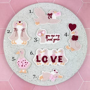 Puede incluir: Una colección de siete adornos decorativos con temática de gansos. Los gansos están en varias poses, algunos sosteniendo corazones, lazos y cartas. La paleta de colores incluye tonos de rosa, blanco y marrón. La palabra "LOVE" está escrita con gansos.
