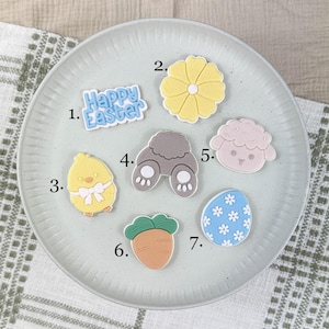 May include: A selection of Easter-themed charms displayed on a plate. The charms feature a yellow chick, a carrot, a lamb, a flower, an egg with blue flowers, bunny feet, and a charm with the text "Happy Easter".