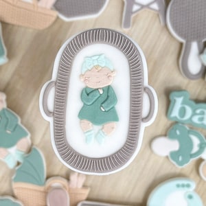 Op de afbeelding: Een set van 15 groene en bruine babyborrelkoekjes, versierd met verschillende babythema-ontwerpen, waaronder een baby in een wieg, een rammelaar, een babyfles en een speen. De koekjes zijn gerangschikt op een houten dienblad.