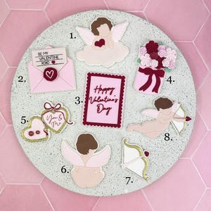 Puede incluir: Colección de decoraciones temáticas de San Valentín sobre una superficie redonda y texturizada. Las decoraciones incluyen querubines, un ramo de flores, un sobre en forma de corazón y un letrero que dice "Happy Valentine's Day". La paleta de colores es principalmente rosa y blanco.