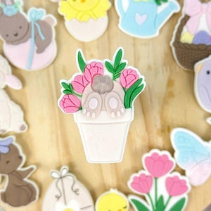 Könnte beinhalten: Ein Holztablett mit verschiedenen Oster-Ausstechformen. Die Ausstechformen haben verschiedene Formen, darunter ein Häschen, ein Küken, eine Blume, ein Schmetterling und ein Ei. Die Ausstechformen sind aus weißem, rosa, blauem und gelbem Fondant.