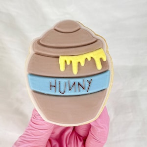 Könnte beinhalten: Ein brauner und gelber Keks, verziert mit einem Honigtopf und dem Wort "Hunny" in blauer Glasur.