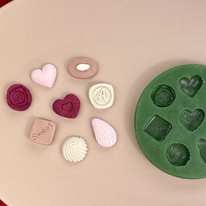 May include: A green silicone mold with 10 different shapes for making candy or chocolate. The mold is on a pink plate with several small, colorful candies already made.