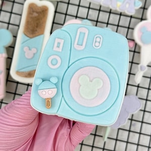 May include: A blue and white decorated sugar cookie shaped like an instant camera with a Mickey Mouse head on the front. The cookie is decorated with pastel colors and has a small Mickey Mouse head lollipop on the side.