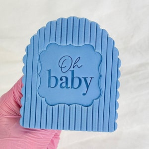 May include: A light blue silicone baby shower sign with the text "Oh baby" in a white frame.