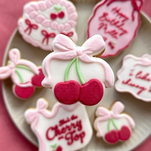 Puede incluir: Un plato de galletas de azúcar decoradas en rosa y blanco con diseños de cerezas y lazos. Las galletas tienen varios mensajes, entre ellos "Happy Birthday Millie", "I like you cherry much" y "Just a Cherry Top".