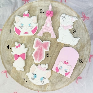 Peut inclure: Divers objets décoratifs blancs et roses, dont des visages de chat, un nœud, une tour Eiffel et un chien. Les articles sont disposés sur un plateau en bois et sont probablement sur le thème d'un personnage de dessin animé. Le fond est blanc avec des motifs de nœuds roses.