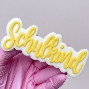 May include: A white, rectangular-shaped plaque with rounded edges featuring the word Schulkind written in raised, yellow, cursive script lettering. The object is held against a plain, light-colored background, showcasing the contrast between the yellow text and the white base.
