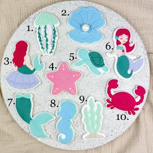 Könnte beinhalten: Eine Sammlung farbenfroher Verzierungen zum Thema Meer, darunter eine Qualle, eine Muschel, eine Meerjungfrau, ein Seestern, eine Schildkröte, eine Krabbe, ein Seepferdchen und Algen. Die Verzierungen sind in Blau-, Rosa-, Grün- und Rottönen gehalten.