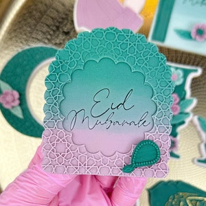Könnte beinhalten: Ein grün und rosa verzierter Keks mit dem Text "Eid Mubarak" in weißer Kursivschrift. Der Keks ist in Form eines Torbogens mit komplizierten Designs.