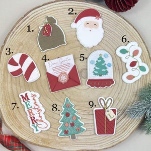 Peut inclure: Une collection d'autocollants découpés sur le thème de Noël sur une surface en bois. Les autocollants comprennent un visage de Père Noël, une canne en bonbon, un sapin de Noël, un cadeau, une boule à neige et une lettre "Cher Père Noël".
