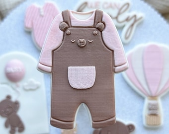 May include: A set of brown and pink fondant cookies decorated with baby-themed designs. The cookies include a baby onesie, a hot air balloon, a teddy bear, and a tag that says "baby".