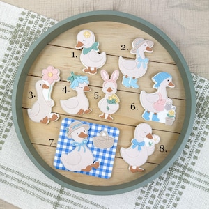 Könnte beinhalten: Eine Sammlung von acht dekorativen Gänsedekorationen, angeordnet auf einem Holztablett. Jede Gans ist in einer einzigartigen Pose dargestellt und trägt Accessoires wie Hüte, Blumen und Schleifen. Die Farbpalette umfasst sanfte Pastelltöne und Erdtöne, wodurch eine charmante, frühlingshafte Ästhetik entsteht.