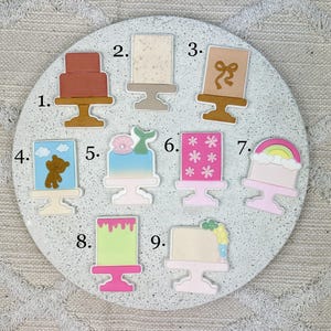Könnte beinhalten: Eine Sammlung von neun dekorativen Anhängern in Kuchenform. Jeder Anhänger hat ein einzigartiges Design, darunter ein mehrstöckiger Kuchen, ein Kuchen mit einer Schleife, ein Kuchen mit einem Teddybären und ein Kuchen mit einem Regenbogen. Die Anhänger befinden sich auf einer runden, gesprenkelten Oberfläche.