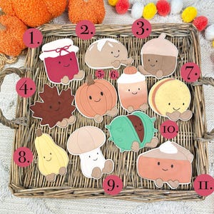 Può includere: Una collezione di ritagli a tema autunnale, in stile cartone animato, disposti su un vassoio intrecciato. I ritagli includono un barattolo di marmellata, una pagnotta, una ghianda, una foglia d'acero, una zucca, un latte, un macaron, una zucca, un fungo, una castagna e una fetta di torta.