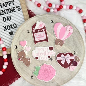 Puede incluir: Una colección de decoraciones temáticas del Día de San Valentín. Incluye un buzón, un globo aerostático, un oso de peluche con globos, un letrero en forma de corazón con el texto "Happy Valentine's Day", una caja en forma de corazón y una rosa rosa. También hay un letrero con el texto "Happy Valentine's Day XOXO".