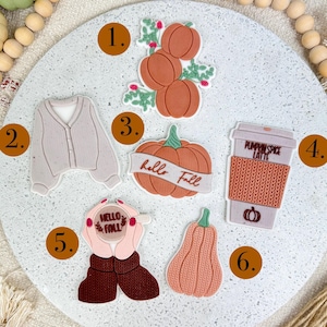 Könnte beinhalten: Eine Sammlung herbstlicher Dekorationen. Das Bild zeigt einen Kürbis, eine Strickjacke, eine Kaffeetasse mit dem Text "Pumpkin Spice Latte" und andere herbstlich inspirierte Designs. Die Farbpalette umfasst warme Töne von Orange, Braun und Beige.