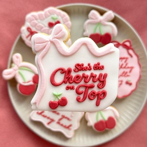 Può includere: Un set di biscotti decorati in rosa e rosso con un tema di ciliegie. Un biscotto ha il testo "She's the Cherry on Top".