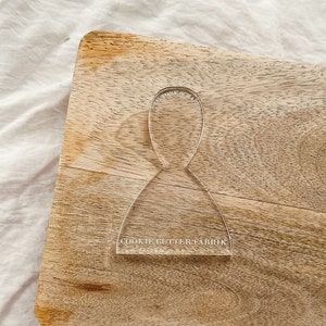 以下が含まれることがあります： 女性のドレスの形をした透明なプラスチック製のクッキーカッター。カッターは木製の表面に置かれ、"COOKIE CUTTER FABRIC"というテキストが印刷されています。