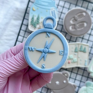 Può includere: Una collezione di biscotti a tema campeggio, tra cui una bussola, un barattolo con alberi, impronte, una mappa, una lanterna, un orso e uno zaino. Il biscotto a forma di bussola ha un bordo azzurro e una rosa dei venti blu.
