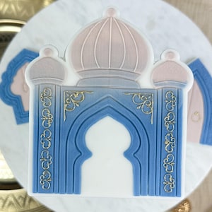 Può includere: Un topper per torta blu e rosa a forma di moschea con accenti dorati. Il topper presenta un tetto a cupola, porte ad arco e dettagli intricati.