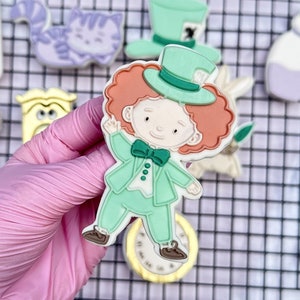 May include: A set of Alice in Wonderland themed sugar cookies, including the Mad Hatter, the Queen of Hearts, a teapot, a cat, and a bottle. The cookies are decorated with colorful icing and details.
