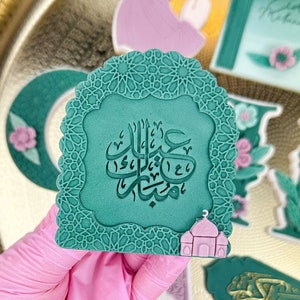 Könnte beinhalten: Ein grüner und weißer Dekor-Keks mit filigranen Mustern und arabischer Kalligraphie. Der Keks ist in Form einer Moschee geformt und hat eine kleine rosafarbene Moschee unten. Der Keks befindet sich auf einem goldenen Tablett mit anderen Dekor-Keksen.