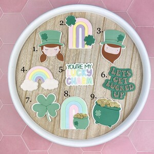 May include: A selection of St. Patrick's Day decorations, featuring a leprechaun face, rainbows, shamrocks, and a pot of gold. The text "YOU'RE MY LUCKY CHARM" and "LETS GET LUCKED UP" are also present. The items are displayed on a wooden surface.