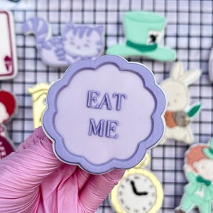 Op de afbeelding: Een verzameling koekjes met het thema Alice in Wonderland. De koekjes bevatten een speelkaart, de Cheshire Cat, de hoed van de Mad Hatter, een fles, de Hartenkoningin, een wit konijn, een klok en een koekje met de tekst "EAT ME".