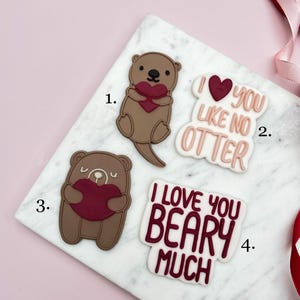 Puede incluir: Cuatro decoraciones temáticas de San Valentín. Uno es una nutria sosteniendo un corazón, otro dice "I love you like no otter", un oso sosteniendo un corazón y otro que dice "I love you beary much".