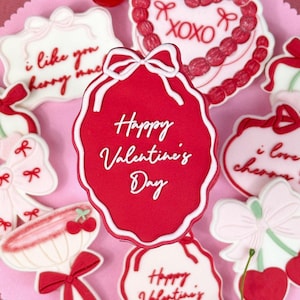 Op de afbeelding: Een roze bord met Valentijnsdag-suikerkoekjes. De koekjes zijn versierd met rode, witte en roze glazuur en hebben verschillende ontwerpen, waaronder harten, strikken en de woorden "Happy Valentine's Day" en "I love you cherry much".