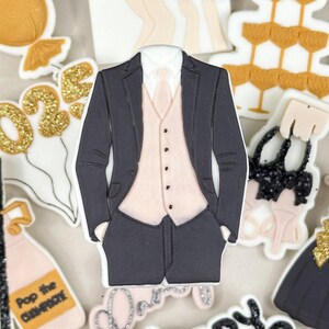 May include: A set of 10 edible cookie cutters for a New Year's Eve party. The cutters are in the shape of a champagne bottle, a champagne glass, a tuxedo, a dress, a bow, a balloon, and the numbers 2023. The cutters are made of white fondant and decorated with gold glitter.