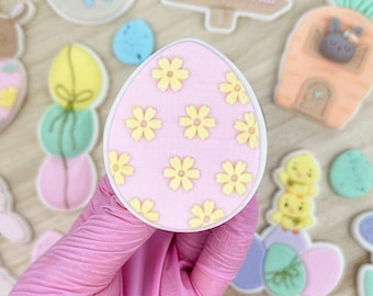 May include: A pink and yellow Easter egg cookie decorated with small yellow flowers. The cookie is being held by a gloved hand. Other Easter-themed cookies are visible in the background, including a bunny, a lamb, a basket of eggs, and a sign that says "Egg Hunt".