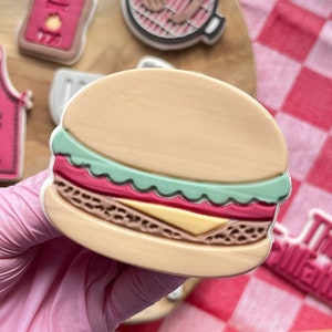 May include: A set of six colourful, decorated sugar cookies with a barbecue theme. The cookies include a hamburger, a grill, a beer mug, a bottle of hot sauce, a king of the grill apron, and a cookie with the text "The Grillfather".