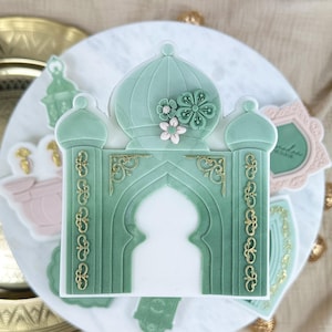 Può includere: Topper per torta decorativo verde e oro con un arco ornato sormontato da una cupola. L'arco è decorato con dettagli dorati e ha un centro bianco. Il topper è circondato da altri elementi decorativi, tra cui una moschea rosa e bianca e una lanterna verde.
