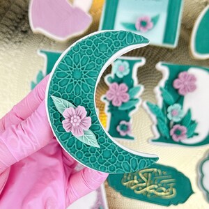 Könnte beinhalten: Ein grüner Halbmond-Keks mit einem weißen und grünen geometrischen Muster und einer rosa Blume mit grünen Blättern. Der Keks liegt auf einem goldenen Tablett mit anderen Keksen, darunter ein grün-weißer Keks mit dem Wort "Eid Mubarak" in arabischer Schrift.