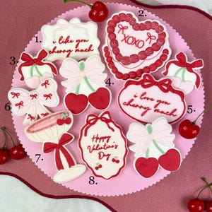 Puede incluir: Una colección de galletas con temática de San Valentín con diseños de cerezas y lazos. Algunas galletas tienen texto que dice "i like you cherry much", "XOXO" y "Happy Valentine's Day". Las galletas están dispuestas sobre una superficie rosa.