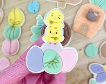 May include: A set of three pastel-colored Easter cookies decorated with a stack of three yellow chicks, a green and white egg with a gold ribbon, and a pink egg. The cookies are on a wooden surface with other Easter-themed cookies.